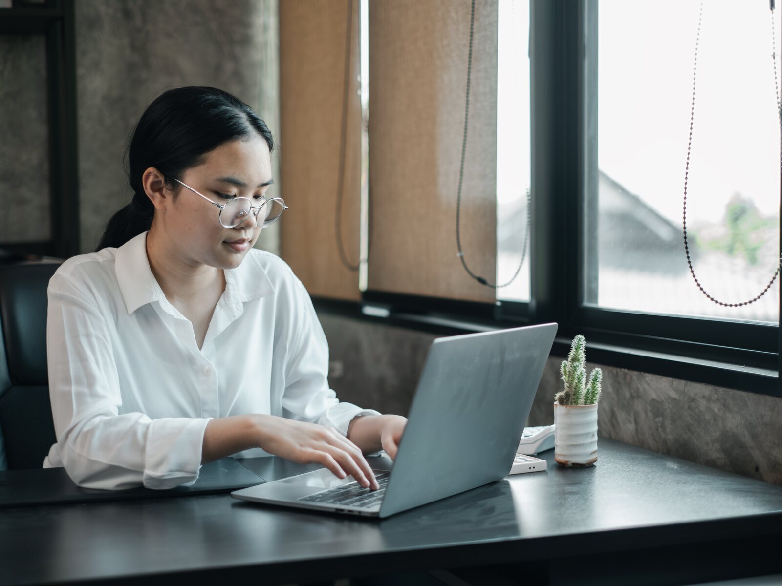 A woman in glasses working at a laptop by a window