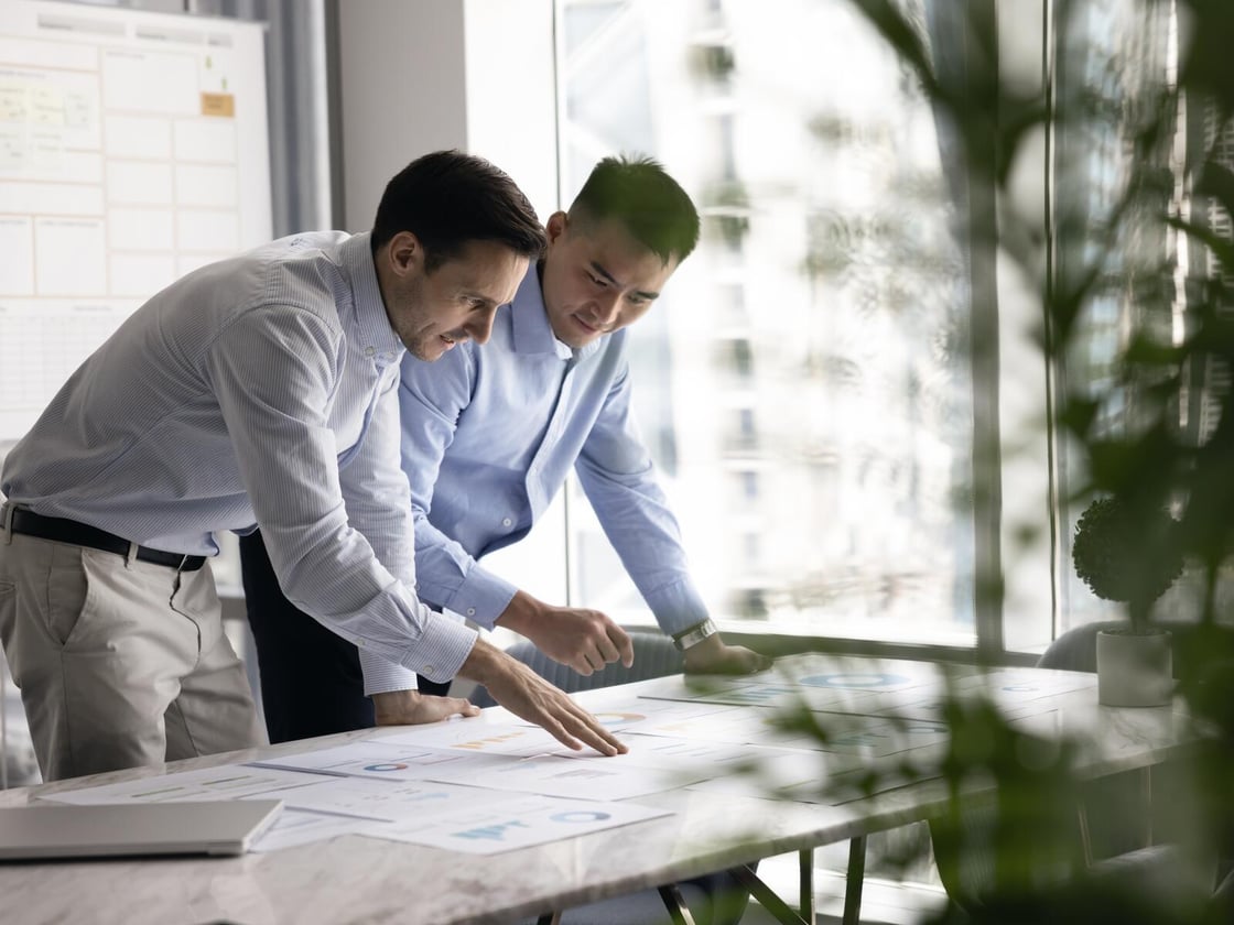 Two colleagues leaning over a table of printed charts, one pointing at a figure.