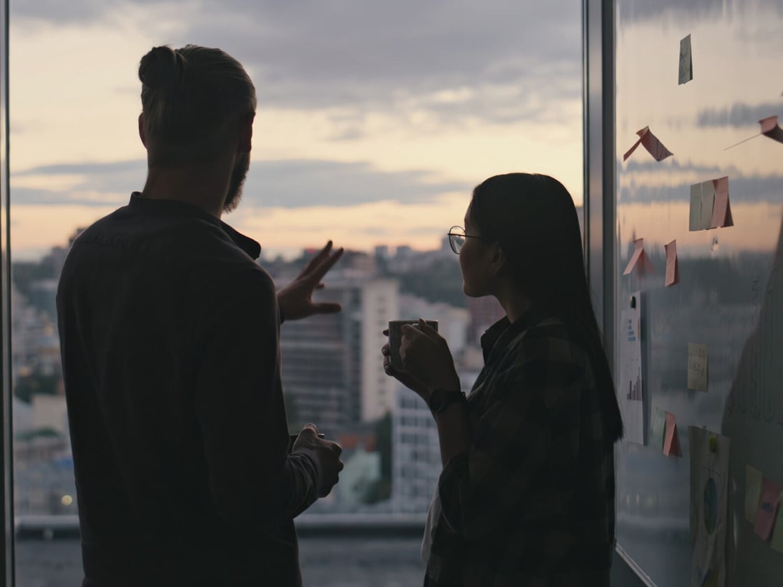 Two colleagues talking at a window at dusk, beside a wall of planning notes and charts.