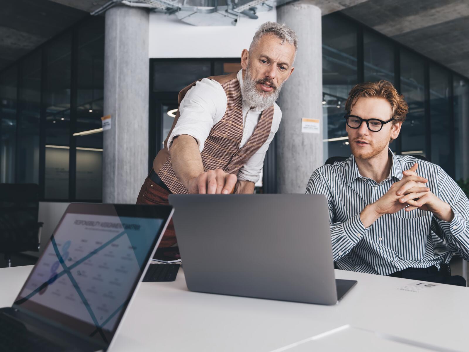 A mentor leans in to point out a detail on a chart to a seated colleague