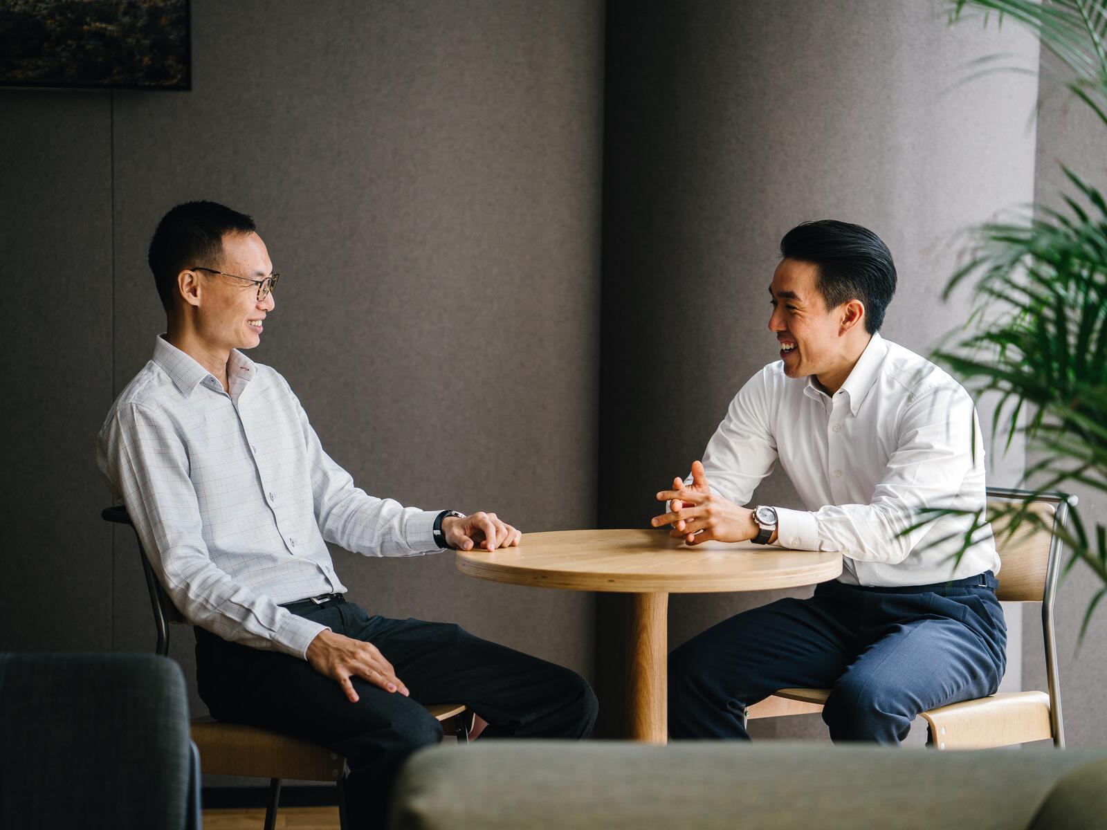 Two colleagues in an animated working conversation across a desk, one talking through a plan