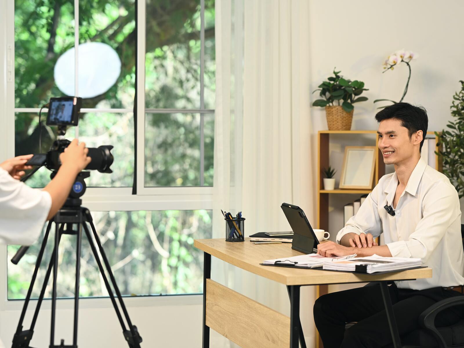 A videographer filming an on-site interview with a woman at her own desk